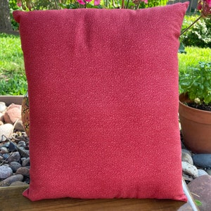 May include: A square decorative pillow with a red speckled pattern. The pillow is placed on a wooden surface, with pink flowers and greenery in the background. The pillow is approximately 16 inches square.