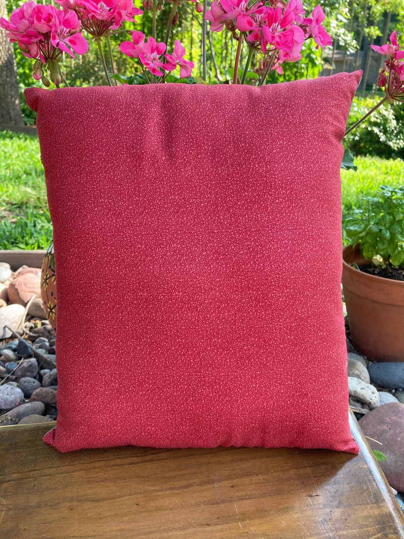 May include: A square decorative pillow with a red speckled pattern. The pillow is placed on a wooden surface, with pink flowers and greenery in the background. The pillow is approximately 16 inches square.