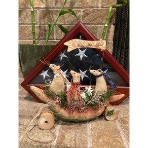 May include: A patriotic display featuring three felt mice in a watermelon boat, adorned with scarves. A banner reading "United We Stand" is displayed in front of a framed American flag. The display includes a wooden spool and decorative moss.