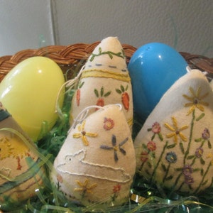 May include: A wicker basket filled with Easter decorations. The basket contains several fabric egg ornaments with embroidered designs, including flowers, carrots, and chicks. Also included are two plastic eggs, one yellow and one blue, and green decorative grass.