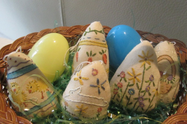 May include: A wicker basket filled with Easter decorations. The basket contains several fabric egg ornaments with embroidered designs, including flowers, carrots, and chicks. Also included are two plastic eggs, one yellow and one blue, and green decorative grass.