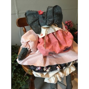 May include: A handmade elephant doll with large, dark gray ears and a pink flower. The doll wears a pink and white checkered dress with a pink skirt and a black band with butterfly designs. The doll is seated on a wooden chair.