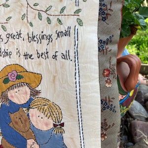 May include: A decorative pillow with a stitched design. The pillow features a drawing of two girls hugging a cat, with the text "is great, blessings small, friendship is the best of all." The pillow has a floral patterned border and a cream-colored background.