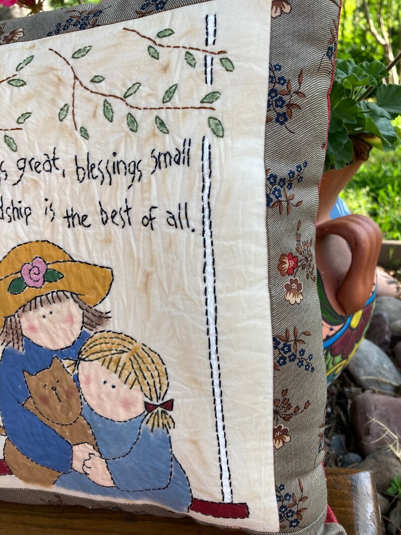 May include: A decorative pillow with a stitched design. The pillow features a drawing of two girls hugging a cat, with the text "is great, blessings small, friendship is the best of all." The pillow has a floral patterned border and a cream-colored background.