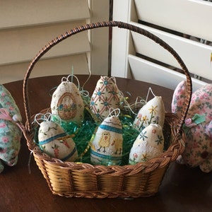 May include: A woven basket filled with Easter egg ornaments and green decorative grass. Two fabric bunnies with floral patterns flank the basket. The eggs are decorated with embroidered designs and hanging loops.
