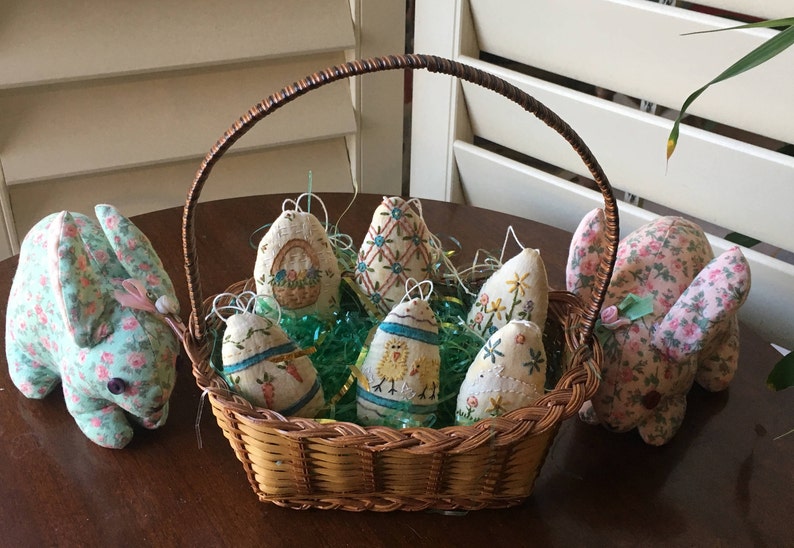 May include: A woven basket filled with Easter egg ornaments and green decorative grass. Two fabric bunnies with floral patterns flank the basket. The eggs are decorated with embroidered designs and hanging loops.