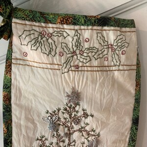 Puede incluir: Un calcet&iacute;n navide&ntilde;o con un frente color crema, bordado con un &aacute;rbol de Navidad y acebo. La parte superior y el ribete son verdes con un estampado de pi&ntilde;as. Los dijes plateados de un copo de nieve, un &aacute;ngel y adornos adornan el &aacute;rbol.