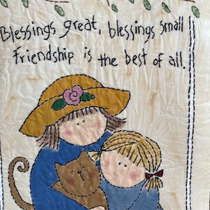 May include: A fabric wall hanging with a stitched design. It features two figures in blue clothing, one wearing a yellow hat, hugging a brown cat. The text "Blessings great, blessings small, Friendship is the best of all" is stitched above the figures.