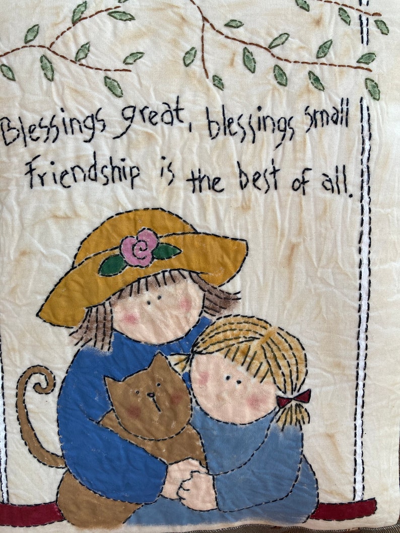 May include: A fabric wall hanging with a stitched design. It features two figures in blue clothing, one wearing a yellow hat, hugging a brown cat. The text "Blessings great, blessings small, Friendship is the best of all" is stitched above the figures.