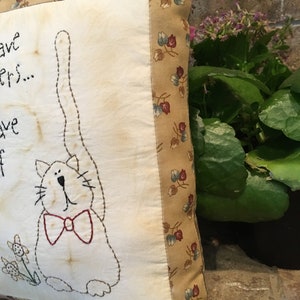 May include: A square, cream-coloured cushion featuring a stitched cat design with a red bow tie. The cushion has a floral patterned border and text that reads "...have...ers...have...off".