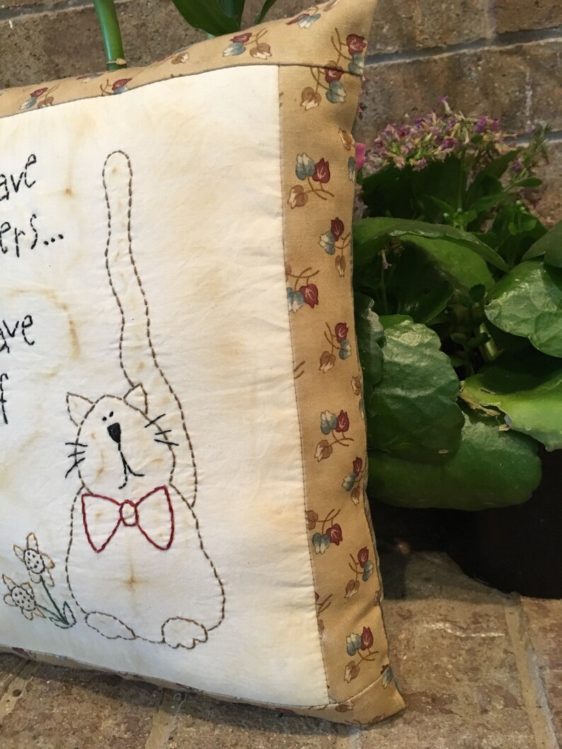 May include: A square, cream-coloured cushion featuring a stitched cat design with a red bow tie. The cushion has a floral patterned border and text that reads "...have...ers...have...off".