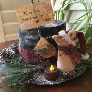 May include: A decorative holiday centerpiece featuring a mouse wearing a Santa hat and scarf, a faux candle, and a sign that reads "I'll be back again some day" Frosty. The centerpiece includes pine needles, berries, and pinecones.