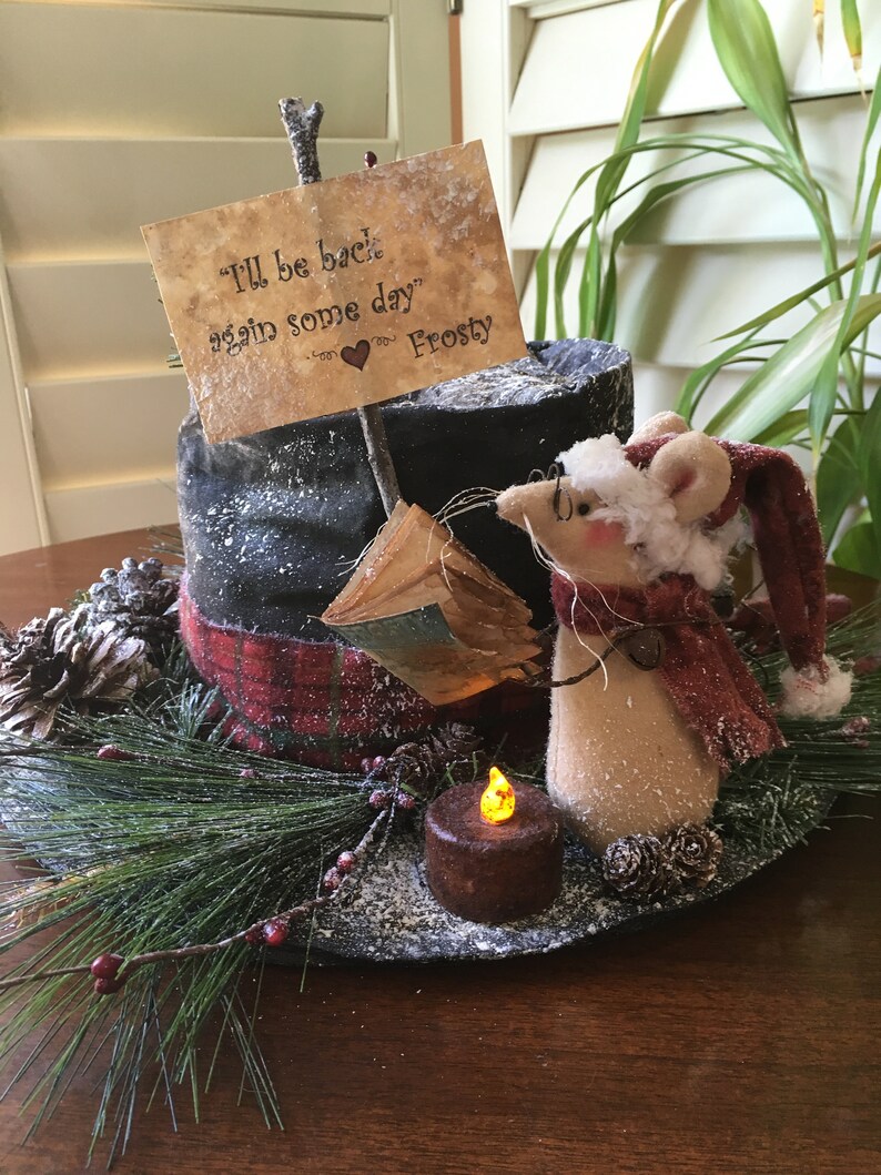 May include: A decorative holiday centerpiece featuring a mouse wearing a Santa hat and scarf, a faux candle, and a sign that reads "I'll be back again some day" Frosty. The centerpiece includes pine needles, berries, and pinecones.