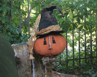 May include: A Halloween decoration featuring an orange pumpkin head with a black witch hat and a burlap scarf. The pumpkin has black eyes and a wooden nose. The decoration is mounted on a rusty spring and sits on a weathered wooden base.