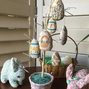 May include: An Easter-themed display featuring a decorative tree adorned with embroidered egg ornaments. Two plush bunny figures with floral patterns flank the tree. A woven basket holds additional Easter eggs, creating a festive scene.