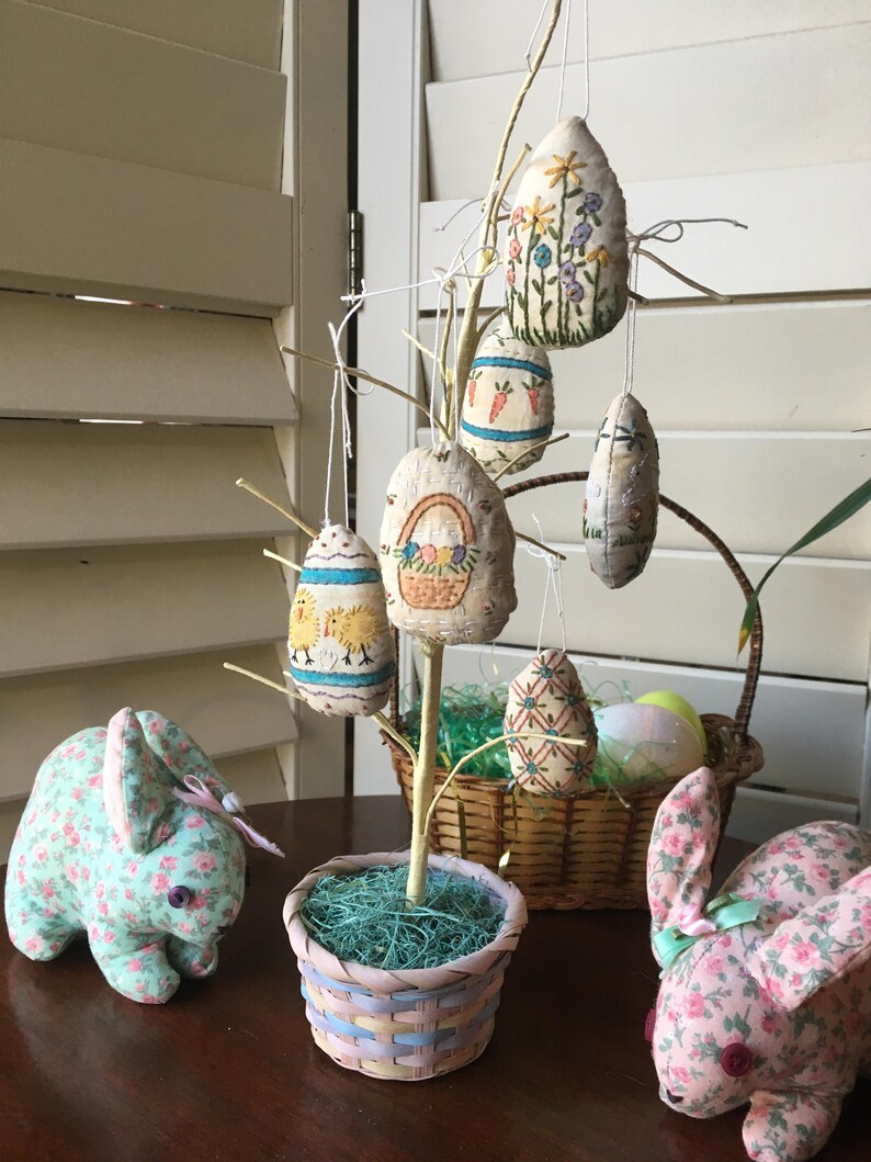 May include: An Easter-themed display featuring a decorative tree adorned with embroidered egg ornaments. Two plush bunny figures with floral patterns flank the tree. A woven basket holds additional Easter eggs, creating a festive scene.