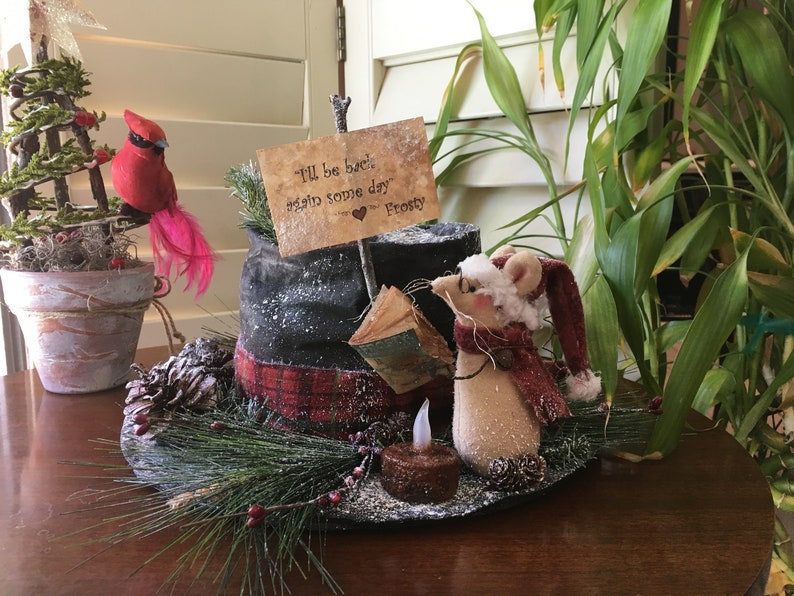 May include: A festive holiday decoration featuring a red cardinal perched on a miniature Christmas tree, a snowman's hat, and a mouse wearing a Santa hat and scarf. The sign reads "I'll be back again some day Frosty."