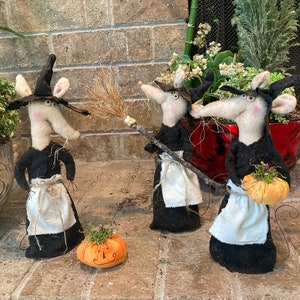 May include: Three whimsical Halloween witch mice figurines. Each mouse wears a black dress, a white apron, and a pointed black hat. One mouse holds a broom, another a pumpkin, and a third stands near a carved pumpkin. The figurines are set on a brick surface.