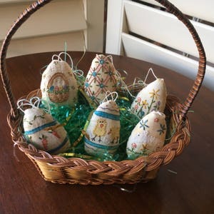 May include: A woven basket filled with six decorative Easter egg ornaments. The eggs are fabric, hand-stitched with floral and other spring-themed designs. The basket sits on a wooden surface, with green and yellow decorative grass.