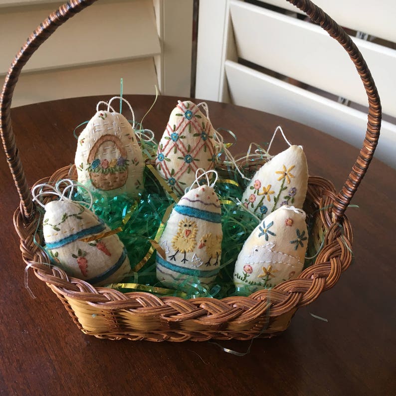 May include: A woven basket filled with six decorative Easter egg ornaments. The eggs are fabric, hand-stitched with floral and other spring-themed designs. The basket sits on a wooden surface, with green and yellow decorative grass.