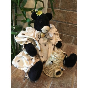 May include: A handmade black bear plush toy wearing a cream-colored dress with a bee pattern and a yellow flower. A decorative beehive made of rope sits nearby. The bear has black paws and a cream-colored face.