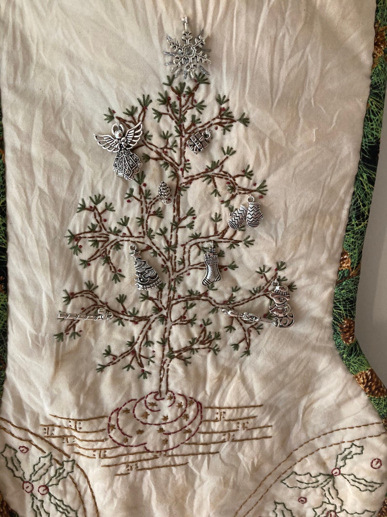 Puede incluir: Calcet&iacute;n navide&ntilde;o color crema con borde estampado en verde y dorado. El calcet&iacute;n presenta un dise&ntilde;o de &aacute;rbol de Navidad bordado adornado con abalorios plateados. El &aacute;rbol est&aacute; decorado con varios adornos, incluyendo un &aacute;ngel, un copo de nieve y un mu&ntilde;eco de nieve.