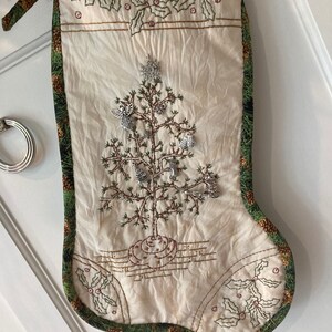 Puede incluir: Calcet&iacute;n navide&ntilde;o color crema con borde verde y marr&oacute;n con pi&ntilde;as. El calcet&iacute;n presenta un &aacute;rbol de Navidad bordado con adornos plateados y un dise&ntilde;o de acebo. Una cinta verde est&aacute; atada en la parte superior.