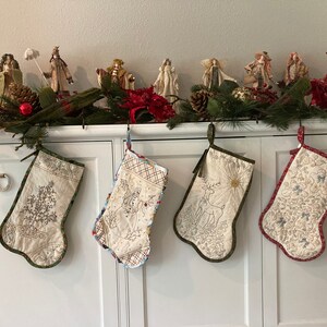 Puede incluir: Cuatro calcetines navide&ntilde;os acolchados con dise&ntilde;os bordados cuelgan de un armario blanco. Los calcetines presentan im&aacute;genes de un &aacute;rbol de Navidad, un mu&ntilde;eco de nieve, un ciervo y motivos florales. Una guirnalda y figuritas decoran la parte superior.