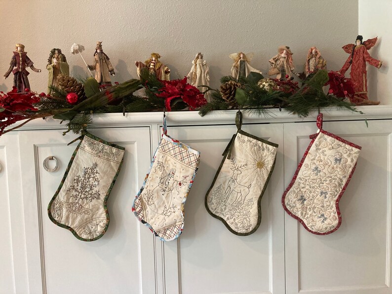 Puede incluir: Cuatro calcetines navide&ntilde;os acolchados con dise&ntilde;os bordados cuelgan de un armario blanco. Los calcetines presentan im&aacute;genes de un &aacute;rbol de Navidad, un mu&ntilde;eco de nieve, un ciervo y motivos florales. Una guirnalda y figuritas decoran la parte superior.