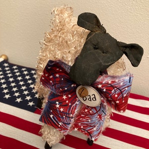 May include: A handcrafted sheep figurine with a black head and curly, cream-colored wool. It wears a red, white, and blue bow with a fireworks pattern and a tag that says "Baa". The sheep is positioned on an American flag.