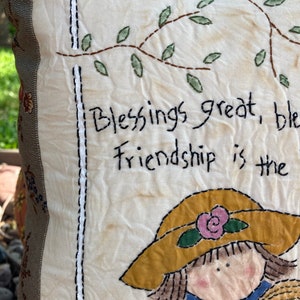 May include: A handmade pillow with a cream-colored front featuring embroidered text and illustrations. The text reads "Blessings great, friendship is the." The illustrations include a girl wearing a yellow hat and a cat. The pillow has a floral patterned border.