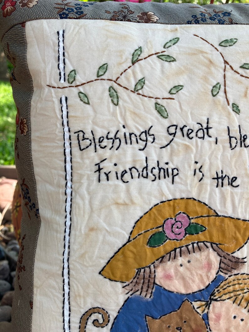 May include: A handmade pillow with a cream-colored front featuring embroidered text and illustrations. The text reads "Blessings great, friendship is the." The illustrations include a girl wearing a yellow hat and a cat. The pillow has a floral patterned border.