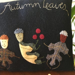 May include: Black rectangular pillow with embroidered text "the Autumn Leaves". The pillow features whimsical leaf and acorn figures in fall colors. The figures are dancing and holding hands. The pillow is on a wooden surface.