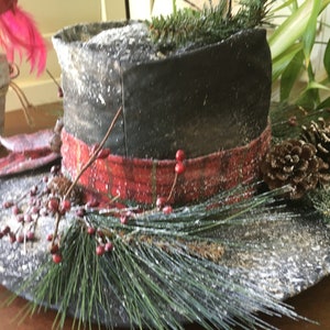 May include: A decorative black top hat with a red band, adorned with pine needles, berries, and pine cones. The hat is dusted with a snowy effect, creating a festive winter theme.