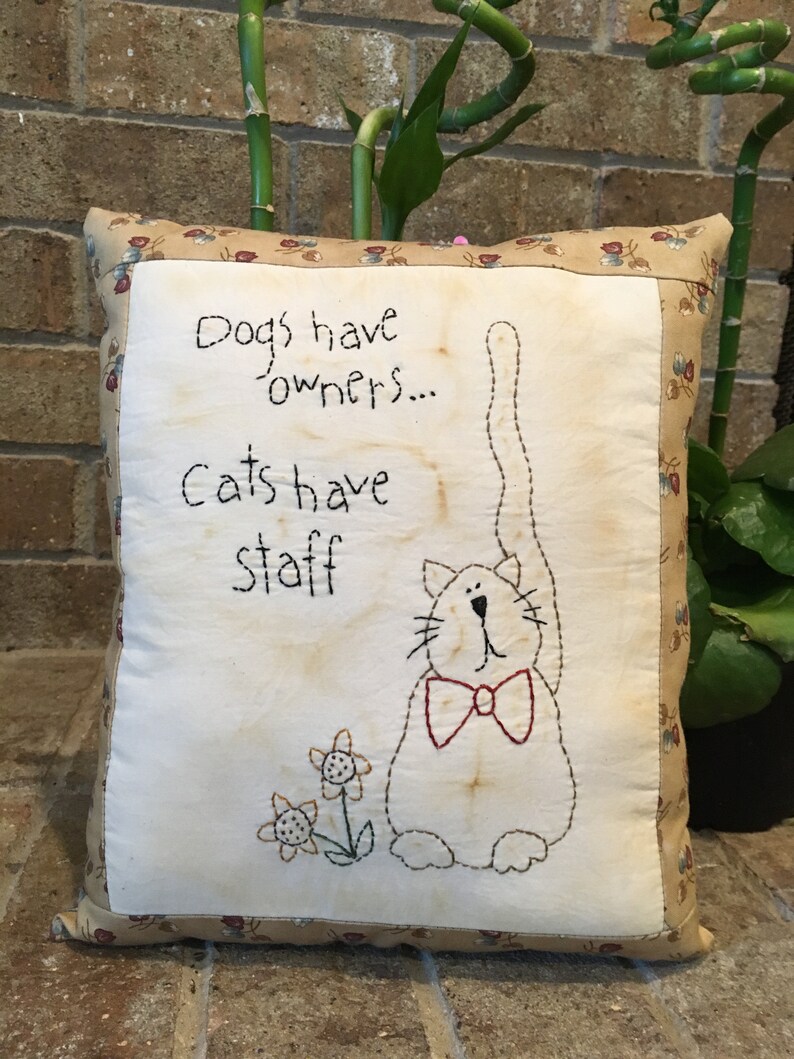 May include: Square cushion featuring a cat design and the text "Dogs have owners... Cats have staff." The cat wears a red bow tie and is next to embroidered flowers. The pillow has a floral border.