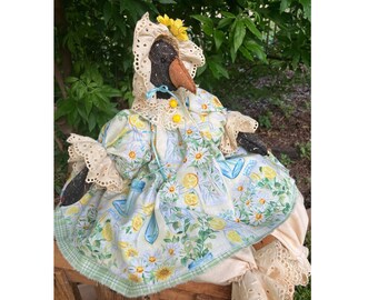 May include: A handmade folk art doll with a black face, a brown beak, and a yellow flower on its head. The doll wears a light blue and white dress with a lemon and daisy print, and a white lace bonnet. The doll is sitting on a wooden surface.