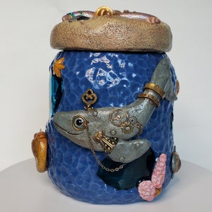 Puede incluir: Un tarro de cerámica azul con una superficie texturizada, decorado con una ballena steampunk plateada y dorada, engranajes y una llave. La tapa es marrón y tiene una superficie texturizada.