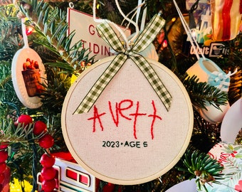 May include: A white, round, embroidered ornament with a green and white checkered ribbon. The ornament says "AVRI" in red thread and "2023 - AGE 5" in black thread.