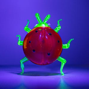 Könnte beinhalten: Eine skurrile Glas-Marienkäfer-Figur. Der Marienkäfer hat einen roten Körper mit schwarzen Punkten und leuchtend grünen Beinen und Fühlern. Der Hintergrund ist ein Farbverlauf aus Lila und Blau.