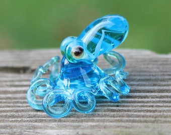 Puede incluir: Una pequeña figura de pulpo de vidrio azul con ocho tentáculos. El pulpo está sentado sobre una superficie de madera.