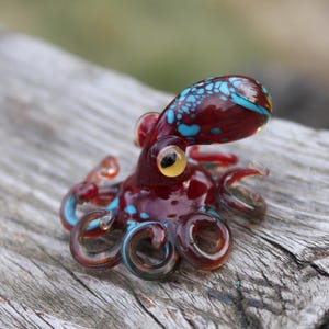 May include: A small glass octopus figurine with a red body and blue and white accents. The octopus has eight tentacles and a large eye.