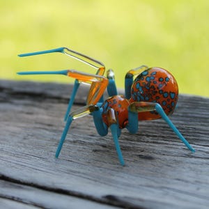 May include: A glass spider sculpture with an orange body speckled with blue dots. The legs and other body parts are a vibrant blue and orange. The sculpture is displayed on a weathered wooden surface.