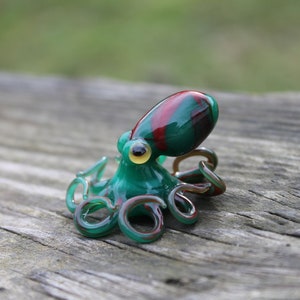 May include: A small glass octopus figurine with a green body and red and black stripes on its head. The octopus has eight tentacles and is sitting on a wooden surface.