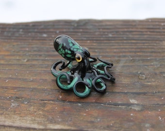 Può includere: Una piccola statuina di polpo in vetro con dettagli neri e verdi. Il polpo ha otto tentacoli e un occhio giallo.