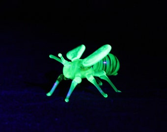 May include: A green glass bee figurine glows under a black light. The bee has a striped abdomen and detailed wings.
