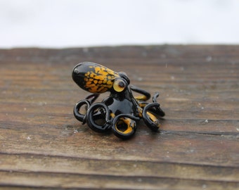 Può includere: Una piccola statuina di polpo in vetro con vetro maculato nero e giallo. Il polpo ha otto tentacoli ed è seduto su una superficie di legno.