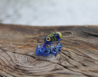 Può includere: Una piccola statuina di polpo di vetro con tentacoli blu e un corpo macchiato di giallo e nero. Il polpo è seduto su un pezzo di legno invecchiato.
