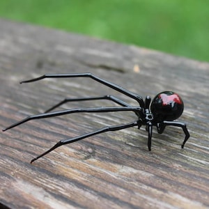 以下が含まれることがあります： 黒いクモの置物で、腹部に赤い模様があります。風化した木製の表面に置かれています。クモは長く細い脚と丸い体を持っています。背景はぼやけた緑色です。