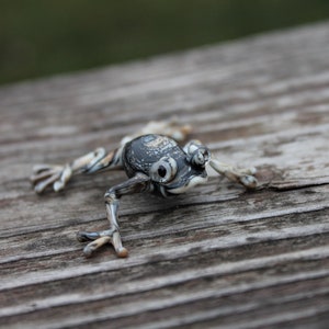 May include: A small, black and white glass frog figurine with detailed eyes and a textured surface. The frog is posed on a wooden surface.