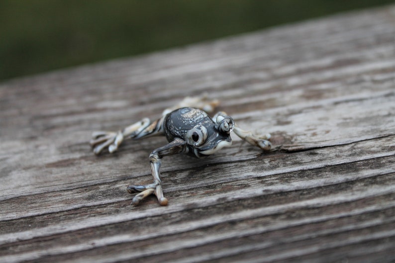 May include: A small, black and white glass frog figurine with detailed eyes and a textured surface. The frog is posed on a wooden surface.